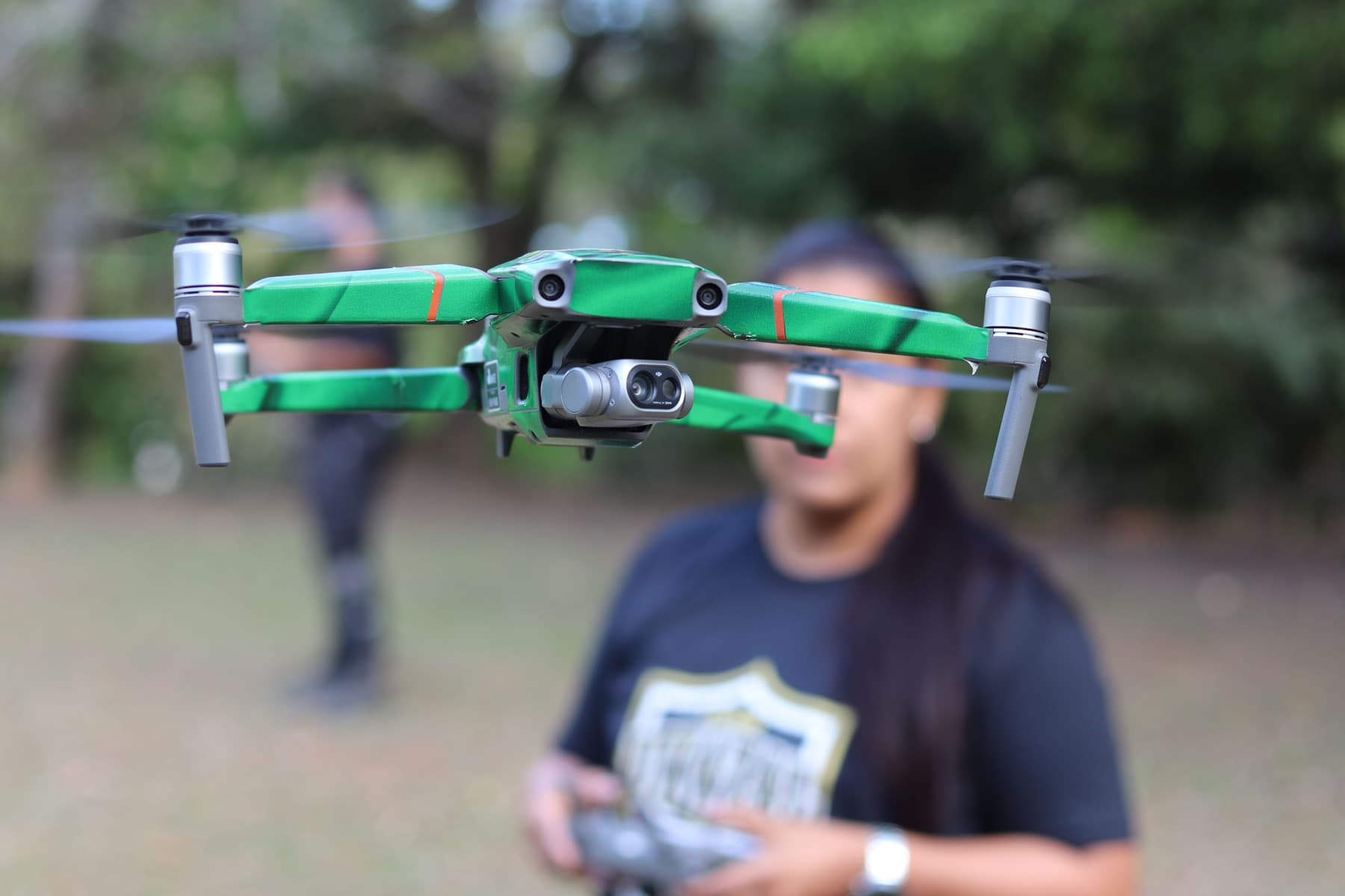 A quadcopter hovering in flight, its pilot on the controller behind it